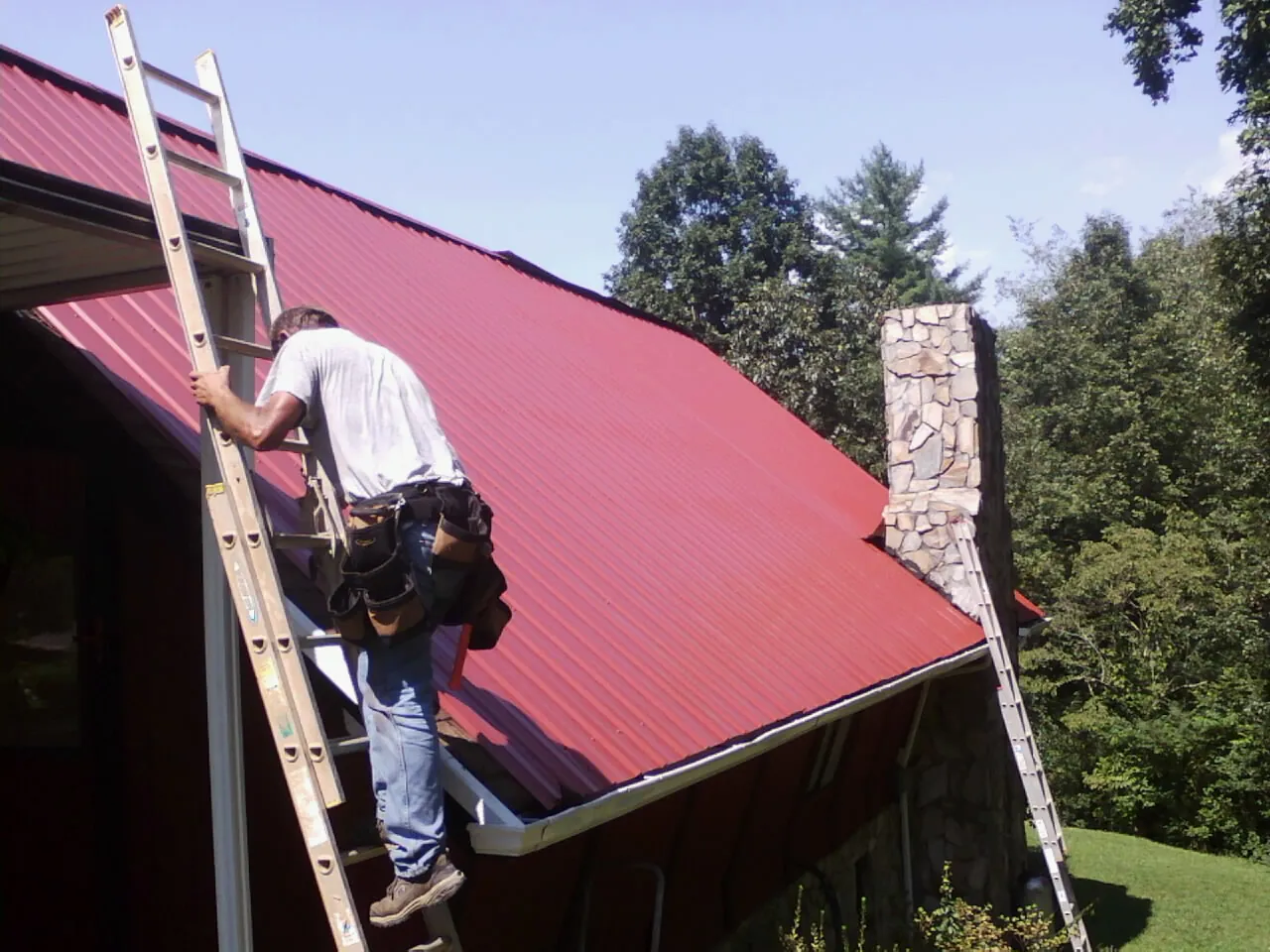 Roofer on a ladder beside a red metal roof and stone chimney — Soffit & Fascia Repair by Roofing Brothers in Boulder City